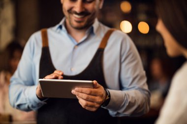 Close-up of happy waiter serving a guest and writing order on digital tablet in a pub. 