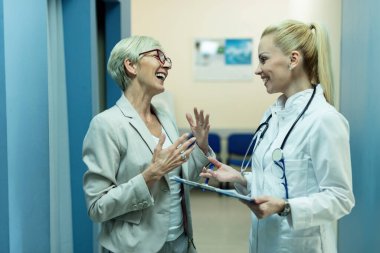 Happy female doctor and mature woman talking while going through medical reports at clinic. 