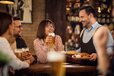 Happy waiter talking with guests who are drinking beer while brining food at their table. 