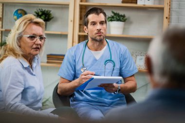 Home caregiver taking notes while communicating with senior patients about their medical condition during a home visit.