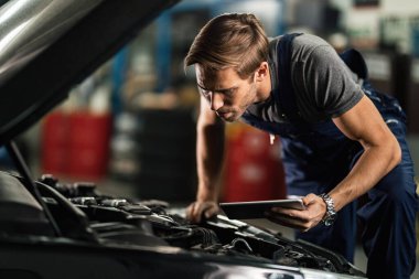 Young auto repairman analzying car engine breakdown while using digital tablet in repair shop. 