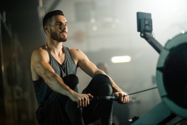 Young athletic man using rowing machine while exercising in health club. 