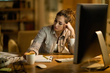 Young displeased woman thinking while drinking tea and working on a computer at night at home. 