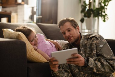 Happy solider and his small daughter relaxing at home and watching something on digital tablet.