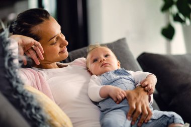 Smiling mother holding her baby boy and looking at him with affection while relaxing on sofa at home. 