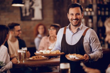 Happy waiter serving food  and looking at camera in a tavern. 