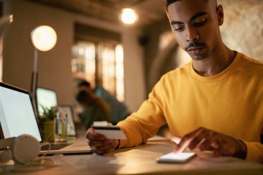 African American businessman using credit cart and mobile phone while checking his online bank account during late night work in the office. 