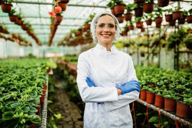 Happy female biologist with crossed arms standing at plant nursery and looking at camera. 