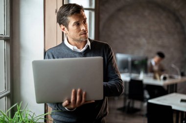 Mid adult entrepreneur thinking of something while using computer by the window in the office.