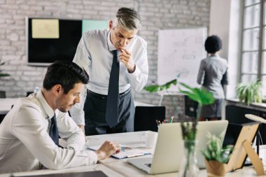 Mid adult businessman thinking while reading business report and analyzing statistics with his younger colleague in the office. 