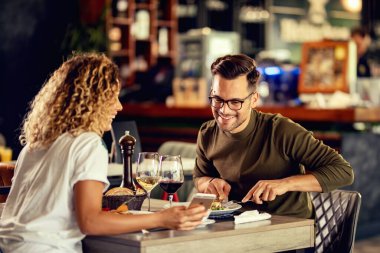 Young happy couple eating in a bar and reading something on smart phone. Focus is on man. 