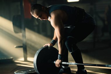 Muscular build man adding weight disc on a barbell while preparing for strength training in a gym. 