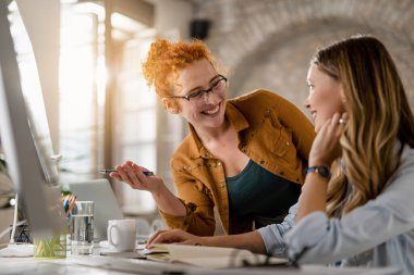 Happy creative businesswomen talking while using desktop PC at casual office. Focus is on redhead woman. 