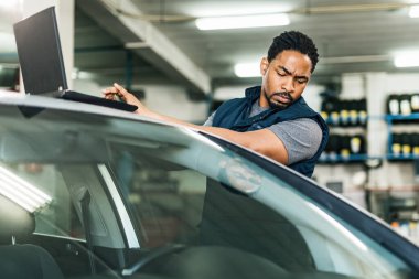 Young black car mechanic working in auto repair shop and using laptop. 