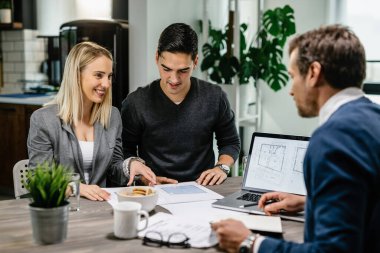Young happy couple communicating with real estate agent while examining blueprints on a meeting. 