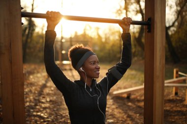 Happy black sportswoman practicing chin-ups at outdoor gym in the park at sunset.  