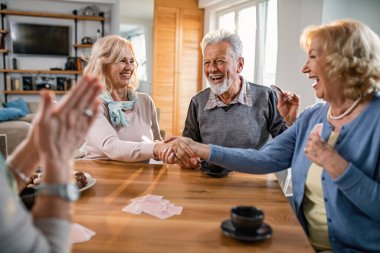 Happy senior women shaking hands and congratulating each other on well playing card game at home. 