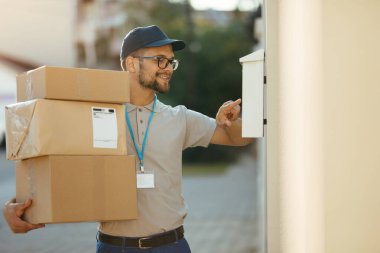 Young happy courier checking name on customer's mailbox while delivering packages at residential district. 