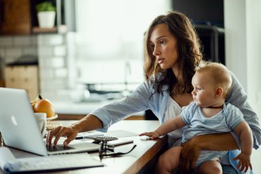 Young woman with baby son working on laptop while planning her home finances. 