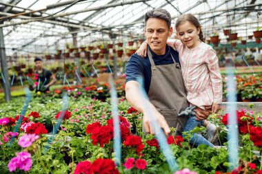 Happy father holding his small daughter and teaching her about type of flowers while working at plant nursery. 