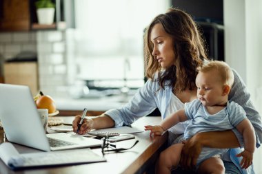 Young mother with baby son working at home and taking notes while planning her finances.