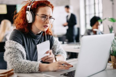 Young casual businesswoman reading problematic e-mail on laptop while listening music on headphones at work. There are people in the background. 