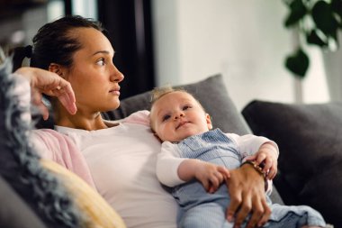 Young mother thinking of something while holding her baby boy and relaxing in the living room. 