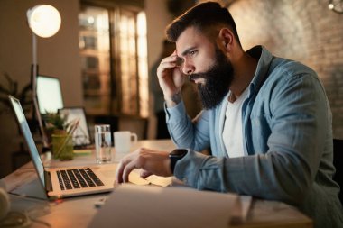 Worried businessman reading an e-mail on a computer while working at night in the office. 