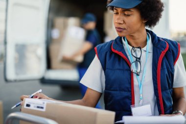 African American worker organizing delivering schedule and checking boxes before the delivery. Her colleague is loading packages in a van in the background. 