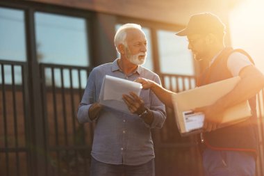 Mature man and deliverer going through paperwork during package delivery. 