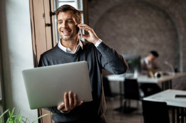 Smiling businessman standing by the window in the office and communicating over cell phone while holding laptop. 