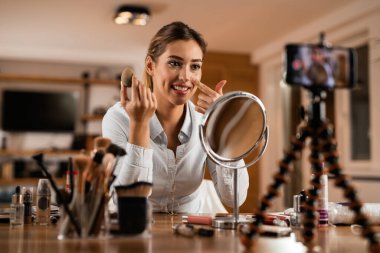 Smiling woman recording make-up vlog while applying face powder at home. 