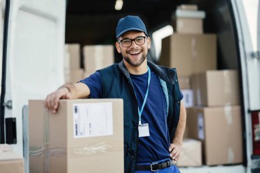 Young happy delivery man loading cardboard boxes in a van and looking at camera. 