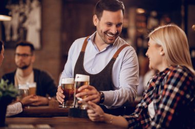 Young happy waiter talking to a woman while giving her a glass of beer in a pub. 