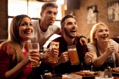Group of cheerful friends having fun while watching sports game on TV and drinking beer in a bar. 