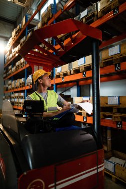 Warehouse worker in a forklift writing notes while checking stock of packages in industrial building. 