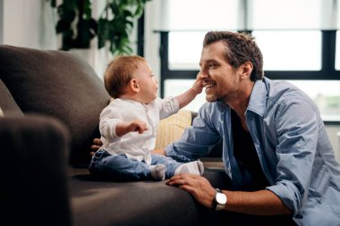 Curious baby son touching father's face while sitting on the sofa in the living room. 