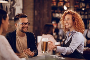 Happy female student working as a waitress and serving beer to a couple in a pub. 