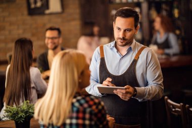 Mid adult waiter using digital tablet and writing an order while communicating with a woman in a bar. 