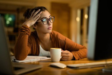 Young pensive woman reading e-mail on desktop PC while working in the evening at home. 