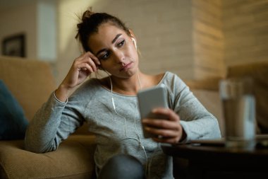 Young pensive woman feeling sad while using smart phone in the evening at home. 