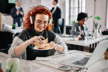 Happy female entrepreneur taking a break from work and eating sandwich in the office. There are people in the background. 