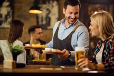 Happy waiter serving tortilla chips to female guest while she drinking beer in a tavern. 
