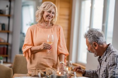 Happy senior woman standing with at dining table with glass of wine while communicating with her husband during lunch time. 