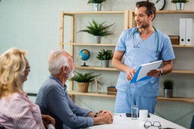 Happy male general practitioner holding medical reports while communicating with mature couple during medical consultations at his office. 