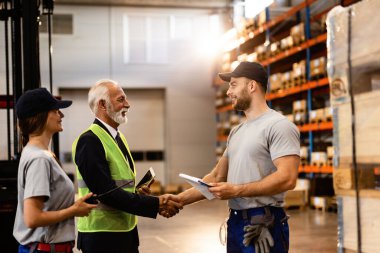 Happy mature businessman handshaking with a worker while visiting factory warehouse. 