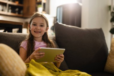 Cute smiling girl using digital tablet in the living room and looking at camera. 