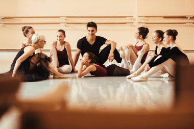 Larger group of ballet dancers and their instructor sitting on the floor and communicating after ballet class at dance studio. 