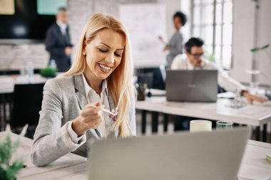 Happy businesswoman eating at her office desk while taking a break from work. There are people in the background. 