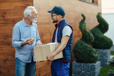 Happy senior man communicating with a courier while signing paperwork for delivered packages. 
